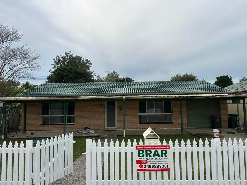 Green tile roof on a brick home with a Brar Roofing sign on the picket fence
